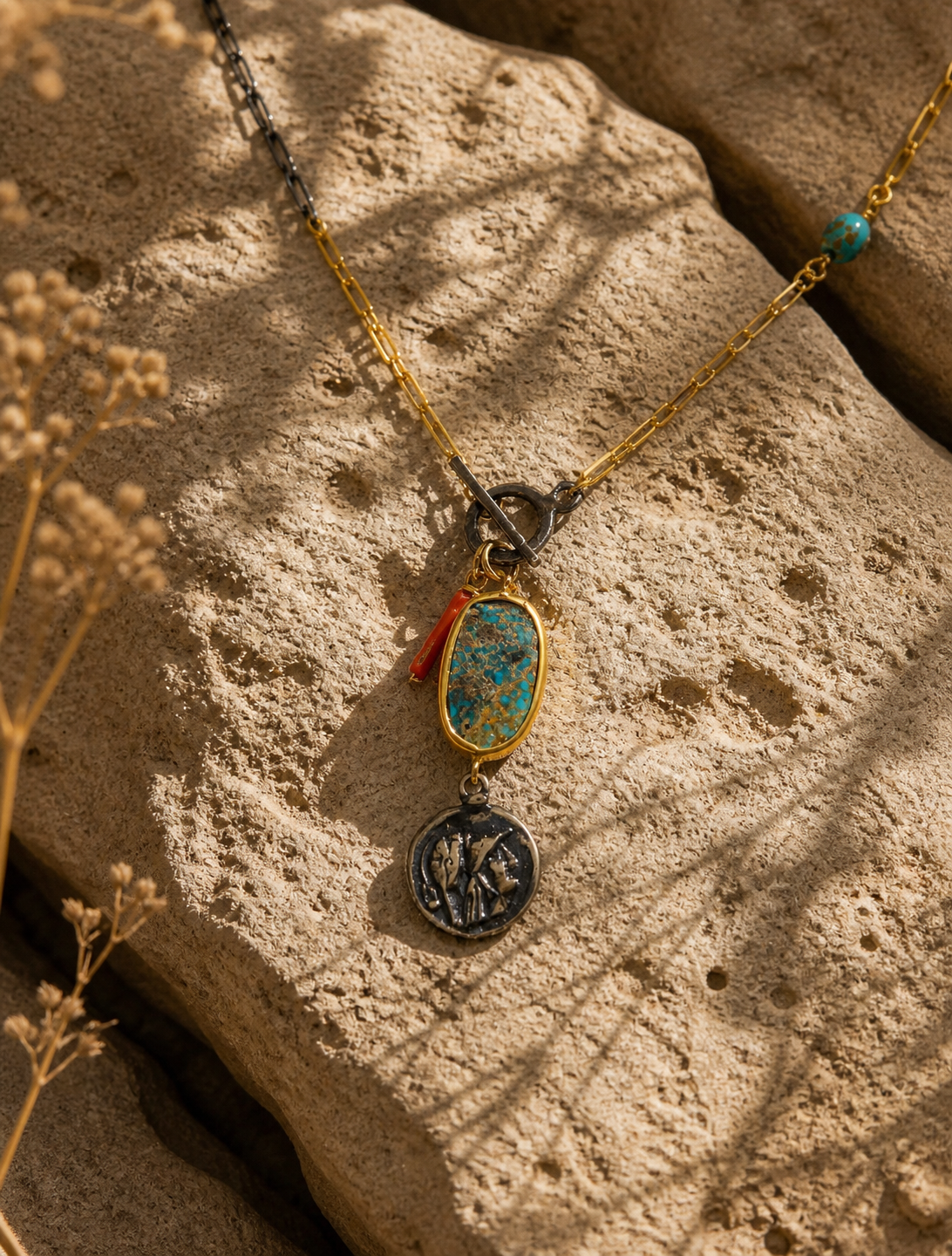 Antique turquoise stone necklace with gold-plated silver chain, red stone charm and oxidized coin motif pendant on natural stone surface